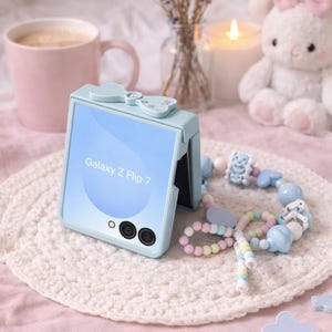 Peut inclure: Un téléphone Galaxy Z Flip 7 bleu clair ouvert est posé sur un tapis crocheté blanc. Le téléphone a un étui bleu clair avec un motif en forme de nœud. Un bracelet de perles aux couleurs pastel et un lapin en peluche sont également dans l'image.
