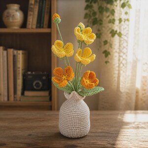 以下が含まれることがあります： 緑の茎と葉を持つ黄色とオレンジ色の花をあしらったかぎ針編みのアレンジメントで、質感のあるオフホワイトのかぎ針編みの花瓶に入っています。花にははっきりとした花びらと中心部があり、花瓶は丸みを帯びた形状をしています。