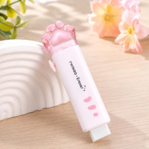 Peut inclure: Une gomme rétractable avec un boîtier en plastique rose clair arborant un motif de patte de chat sur le dessus. Le corps porte l'inscription TWINGOO • Eraser? et trois petits motifs de coussinets roses. La pointe blanche est visible à la base.