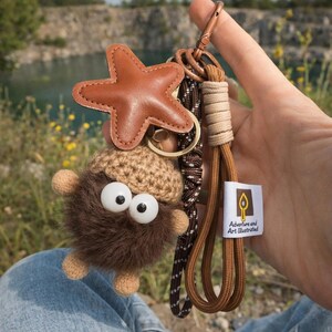 Peut inclure: Un porte-clés artisanal avec un charm en cuir marron en forme d'étoile, une créature en peluche fantaisiste avec de grands yeux globuleux et une sangle tissée. Le porte-clés comprend une étiquette avec l'inscription "Adventure and Art Illustrated."