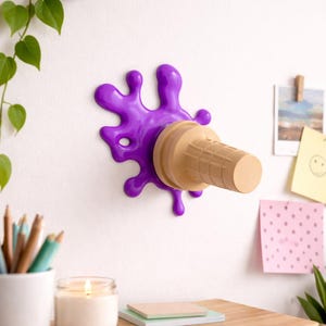 Può includere: Un fantasioso porta-cono gelato da parete. Il supporto presenta un design viola a goccia di vernice con un cono gelato color beige. Il cono è realizzato in materiale a forma di cialda. Il supporto è montato su una parete bianca.