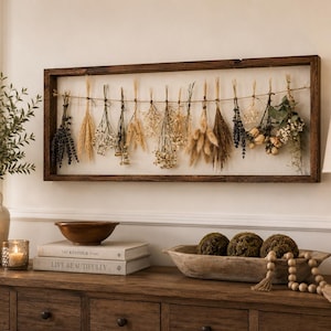 May include: A rectangular dark brown wooden frame containing a horizontal twine string with various dried flowers and grasses hanging from it. The arrangement includes lavender, wheat, baby's breath, and other dried botanical elements in neutral tones.