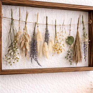 May include: A rectangular wooden frame containing a horizontal twine string from which various dried flowers and grasses are suspended. The arrangement includes white, yellow, purple, and beige botanical elements hanging in a row.