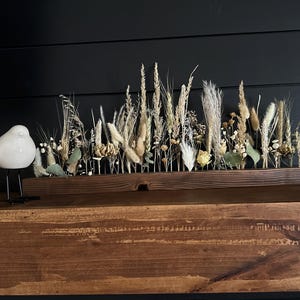 May include: A long, rectangular dark brown wooden base holds an arrangement of dried grasses, wheat stalks, small white flowers, and green eucalyptus leaves. To the left of the wooden base stands a small, smooth, white ceramic bird figurine.