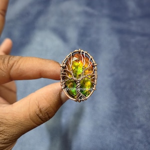 Puede incluir: Anillo ovalado de alambre de cobre con una piedra preciosa colorida. La piedra tiene tonos de amarillo, naranja y verde. Un diseño de árbol está elaborado dentro de la piedra preciosa. El anillo es sostenido por una persona.
