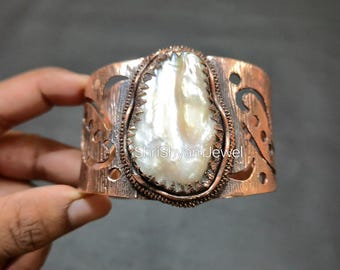 Può includere: Bracciale a polsino in rame con una grande perla barocca bianca. Il bracciale presenta motivi traforati e una finitura strutturata. Il testo "ShriShyamJewel" è visibile.