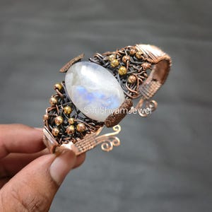 May include: A handcrafted copper cuff bracelet featuring a large, oval moonstone. The bracelet has intricate wirework with gold-colored beads and accents. The text "ShriShyamJewel" is visible on the bracelet.