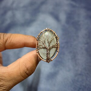 Peut inclure: Pendentif ovale avec un motif d'arbre en fil de cuivre. L'arbre est posé sur une pierre grise. Le pendentif est tenu par une main sur un fond bleu. Le bijou est un pendentif.