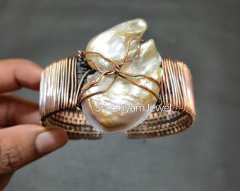 Può includere: Un bracciale a polsino in rame fatto a mano con una grande perla di forma irregolare al centro. La perla è fissata con filo di rame e il polsino stesso è realizzato con filo di rame avvolto strettamente. Il bracciale ha un'estetica naturale e organica.