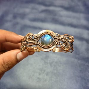 Puede incluir: Un brazalete artesanal de alambre de cobre con una piedra preciosa azul redonda en el centro. El brazalete tiene un intrincado trabajo de alambre con patrones arremolinados y un acabado pulido, mostrando un diseño artístico único. El brazalete es de estilo brazalete.