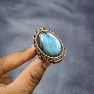 Puede incluir: Anillo ovalado con una gran piedra preciosa azul-verdosa. La piedra está engastada en un marco de alambre de color cobre. El anillo está sostenido por los dedos de una persona sobre un fondo borroso.