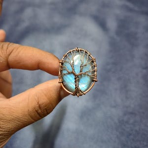 Può includere: Ciondolo ovale con una pietra preziosa blu e un design ad albero avvolto in filo metallico. Il filo color rame forma il tronco e i rami dell'albero, sullo sfondo della pietra blu. Il ciondolo è tenuto in mano.