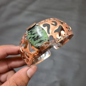 May include: Copper cuff bracelet featuring a large, oval green and black stone. The bracelet has an openwork design with cut-out shapes and the text "ShriShyamJewel". The cuff is held by a hand.