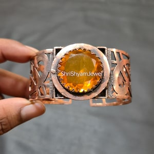 Op de afbeelding: Een koperen manchetarmband met een open ontwerp en een grote, ronde, gele edelsteen in het midden. De armband heeft een rechthoekig frame rond de edelsteen en de tekst "ShriShyamJewel".