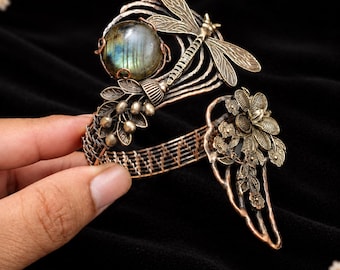 Puede incluir: Un brazalete de alambre de cobre con una piedra preciosa de labradorita iridiscente redonda, un motivo detallado de libélula y un grupo de flores y hojas de metal. La banda está construida con alambre de cobre tejido.
