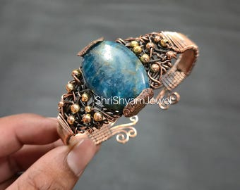 May include: A handcrafted copper cuff bracelet featuring a large, oval, blue gemstone at the center. The bracelet has intricate wirework with small gold-colored beads and the text "ShriShyamJewel".