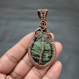 Puede incluir: Colgante envuelto en alambre de cobre con una piedra verde en el centro. El colgante presenta un diseño de árbol y el texto "Shri Shyam Jewel". El trabajo de alambre forma una forma de lágrima, mostrando los patrones naturales de la piedra.