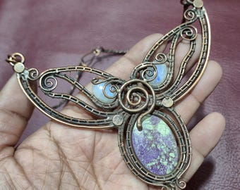 May include: Handmade copper wire necklace featuring a central oval stone with purple and green hues, framed by intricate wirework. The necklace includes two smaller, round, iridescent stones and decorative swirls. The design is held in a hand.