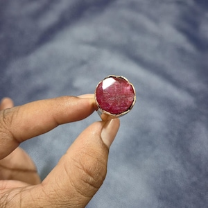 May include: A ruby ring with a large, faceted, oval-shaped red gemstone. The gemstone is set in a gold-colored bezel on a silver band. The ring is held up against a blue background.