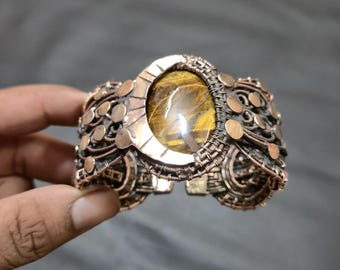 May include: Copper-toned cuff bracelet featuring a large, oval tiger's eye stone at the center. The bracelet has intricate wirework and decorative circular accents. The open-ended design allows for easy wear.