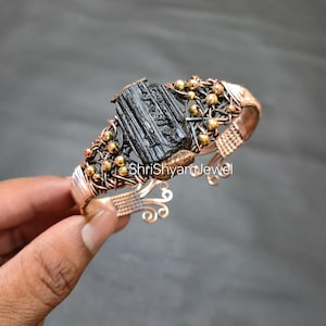 May include: A handcrafted copper wire bracelet featuring a large, raw black tourmaline stone at its center. The bracelet is adorned with small gold-colored beads and intricate wirework. The text "ShriShyam Jewel" is visible on the bracelet.