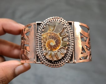 May include: Copper cuff bracelet with an oval fossil centrepiece. The fossil is set within a woven wire frame. The bracelet has cut-out geometric designs on the sides. The text "Shri Shyam Jewel" is visible on the bracelet.