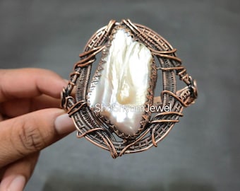 Può includere: Anello artigianale in filo di rame con una grande gemma bianca perlacea irregolare. La gemma è il punto focale, incastonata in una montatura in filo di rame intrecciato. L'anello è tenuto da una mano su sfondo grigio.