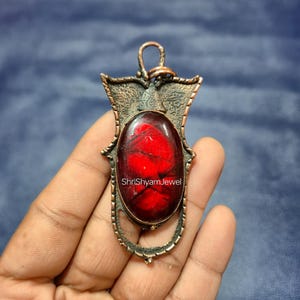 May include: An ornate pendant featuring a large, oval, deep red stone set in a copper-coloured metal frame. The pendant has a loop for a chain and the text "ShriShyamJewel" is visible. The metalwork has intricate detailing.