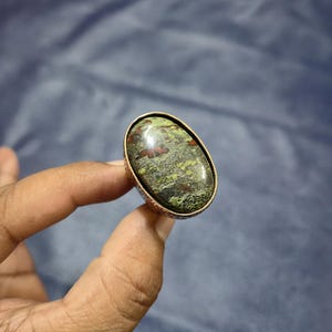 May include: Oval-shaped ring featuring a large, polished stone with a mottled pattern of green, yellow, and red hues. The stone is set in a copper-colored metal band with a textured design. The ring is held up by a hand against a blue background.