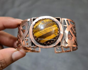 May include: Copper cuff bracelet featuring a large, round tiger's eye stone. The bracelet has an open design with cut-out patterns and the text "ShriShyam Jewel". The tiger's eye stone is a mix of brown and gold.