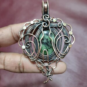 Può includere: Un ciondolo fatto a mano con una pietra verde al centro, racchiuso in un'intricata filigrana di rame. Il ciondolo presenta un design decorativo con volute e accenti circolari, e un'asola per appenderlo. Il metallo ha una finitura calda e antica.