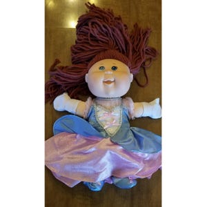 May include: A vintage Cabbage Patch Kids doll with a round face, blue eyes, and a smiling expression. The doll has red yarn hair and is wearing a blue and pink princess dress with matching shoes. The doll's arms are outstretched.