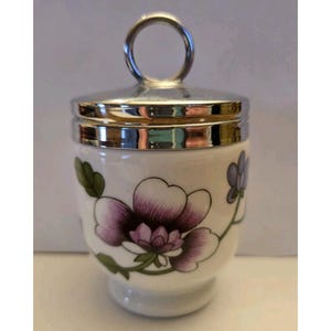 May include: White ceramic egg cup with a silver-tone lid and handle. The cup is decorated with a large purple and white floral design with green leaves. The egg cup is a classic kitchenware item.