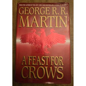 May include: A hardcover book titled "A Feast for Crows" by George R. R. Martin. The cover is predominantly red with gold lettering and a red embossed illustration of a bird. The text "FROM THE AUTHOR OF THE NEW YORK TIMES BESTSELLER A STORM OF SWORDS" is at the top.