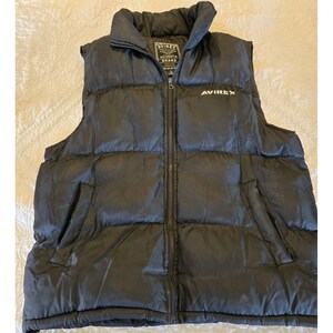 May include: Black Avirex puffer vest with a full-length zipper and two side pockets. The vest has a high collar and the Avirex logo is visible on the chest. The vest is made of a shiny, quilted material.