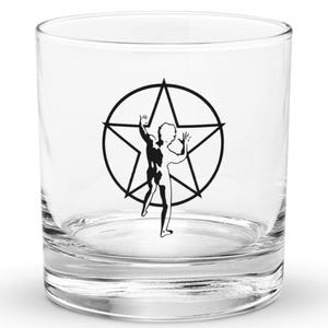 May include: A clear, round tumbler glass featuring a black and white graphic of a person within a star. The glass is empty and rests on a white surface. The design is a stylized illustration.