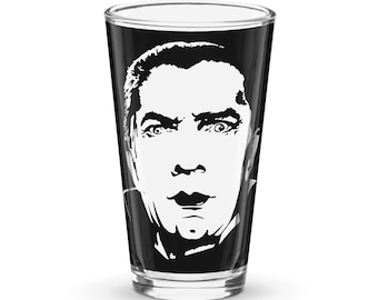 May include: A black pint glass featuring a white graphic of a man's face. The face has a surprised expression with wide eyes and an open mouth. The glass is clear with a thick base.