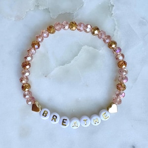 May include: A beaded bracelet featuring a mix of pink and gold faceted beads. The bracelet includes white letter beads spelling "BREATHE" and gold heart-shaped accents. The bracelet is designed for everyday wear.