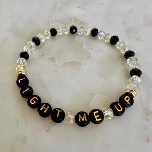 May include: A beaded bracelet with black and clear beads. The bracelet has black beads with gold lettering that spells "LIGHT ME UP". The bracelet is on a white marble surface.