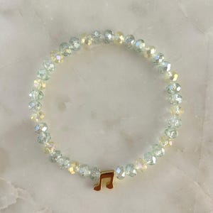 May include: A beaded bracelet featuring iridescent, faceted beads in shades of pale green and yellow. A gold-colored musical note charm is attached to the bracelet. The bracelet is displayed on a white marble surface.