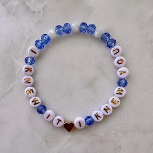 May include: A circular beaded bracelet featuring white round beads with gold lettering that spells out I KNEW IT I KNEW YOU. The design is interspersed with faceted blue crystal beads, small white round beads, and a single gold heart-shaped charm.