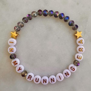 May include: A beaded bracelet featuring iridescent dark gray faceted beads, two gold-toned star charms, and white round beads with gold lettering that spells out the phrase IN MY RUNNING ERA, arranged in a circular pattern on a light surface.