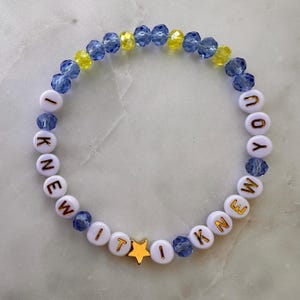 May include: A beaded bracelet featuring alternating blue and yellow faceted crystal beads and white round beads with gold lettering that spells out I KNEW YOU I KNEW IT. A single gold-toned star-shaped bead is positioned near the bottom center.