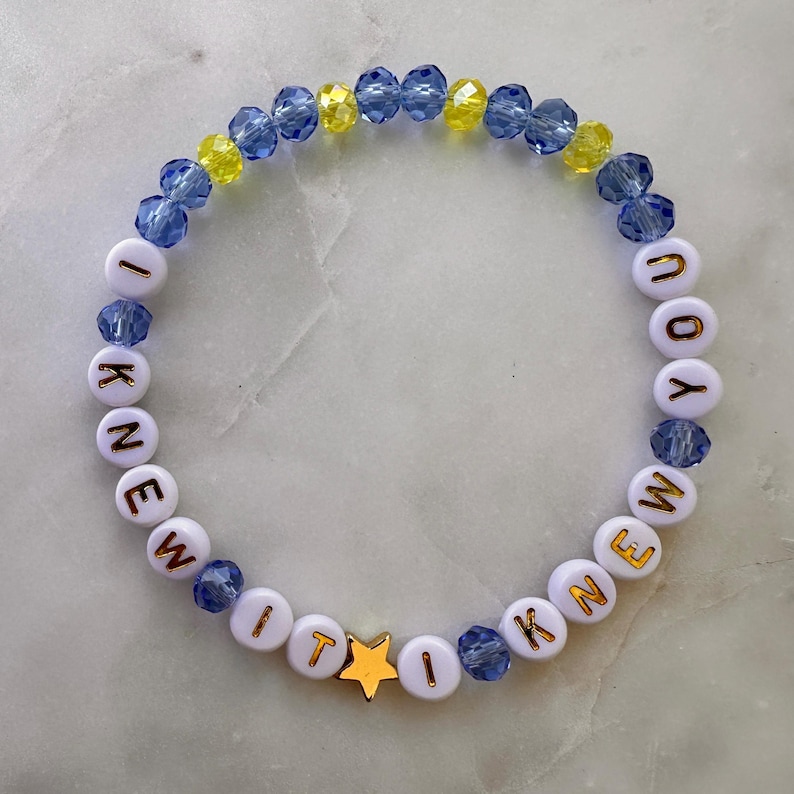 May include: A beaded bracelet featuring alternating blue and yellow faceted crystal beads and white round beads with gold lettering that spells out I KNEW YOU I KNEW IT. A single gold-toned star-shaped bead is positioned near the bottom center.