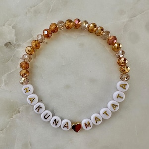 May include: A beaded bracelet with the words "HAKUNA MATATA" spelled out in gold letters on white beads. The bracelet also features a small gold heart and amber-colored crystal beads.