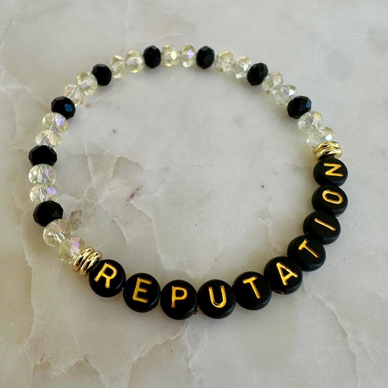 May include: A beaded bracelet with the word "REPUTATION" spelled out in gold letters on black beads. The bracelet also features clear and black beads, with gold accents.