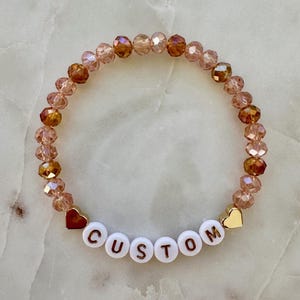 May include: A beaded bracelet with the word "CUSTOM" spelled out in white letter beads. The bracelet features a mix of peach and brown-toned crystal beads, with gold and red heart accents.