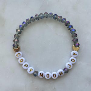 May include: A beaded bracelet with the words "COPY MOON JOY" spelled out in gold letters on white beads. The bracelet features iridescent gray crystal beads and gold crescent moon charms.