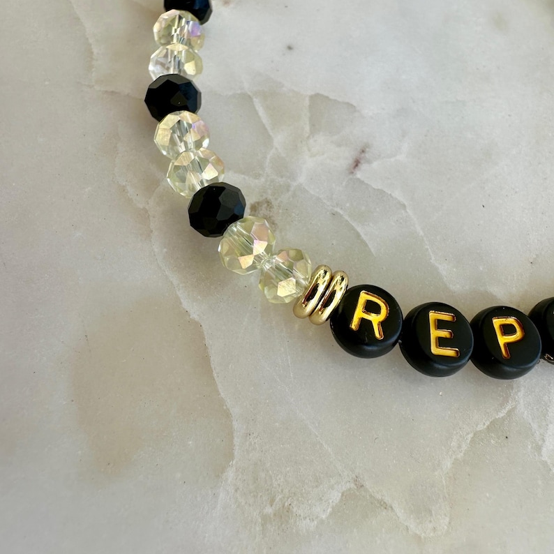 May include: A beaded bracelet featuring black and clear crystal beads, with gold-colored spacers. The bracelet includes black beads with the letters "REP" in gold.