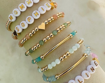 Può includere: Una collezione di braccialetti con perline dorate. Alcuni braccialetti presentano perline con lettere bianche che compongono le parole "MOMMY", "SOPHIE" e "ADELINE". Altri braccialetti hanno dettagli a cuore e a barra dorati e perline di pietre preziose colorate.